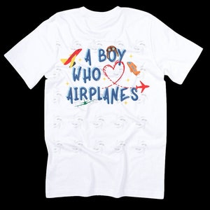 Pu&ograve; includere: Maglietta bianca con una grafica blu e rossa che dice "A Boy Who Loves Airplanes". La grafica include un cuore con ali di aeroplano e un cappello da pilota.