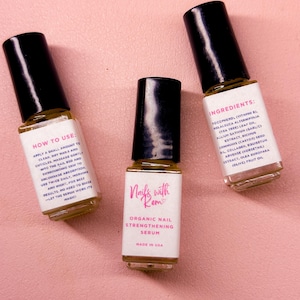 Puede incluir: Tres peque&ntilde;os frascos de s&eacute;rum para u&ntilde;as con tapas negras y etiquetas blancas. Las etiquetas dicen "Nails with Rem" y "Organic Nail Strengthening Serum". Un frasco enumera los ingredientes, otro proporciona instrucciones de uso. El fondo es una superficie rosa claro.