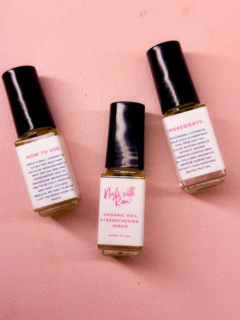 Puede incluir: Tres peque&ntilde;os frascos de s&eacute;rum para u&ntilde;as con tapas negras y etiquetas blancas. Las etiquetas dicen "Nails with Rem" y "Organic Nail Strengthening Serum". Un frasco enumera los ingredientes, otro proporciona instrucciones de uso. El fondo es una superficie rosa claro.