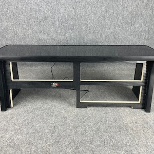 May include: A black wooden shelf unit with two shelves. The shelves are made of a carbon fibre material with a white trim. The top shelf is flat and the bottom shelf has a slight curve.