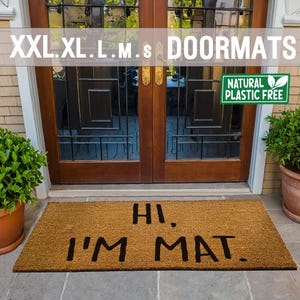 Puede incluir: Una alfombra de coco marrón con el texto "HI. I'M MAT." en letras negras. La alfombra está delante de una puerta de madera con paneles de vidrio. Una planta verde está en una maceta de terracota a la derecha de la alfombra. Un cartel verde "NATURAL PLASTIC FREE" está en la puerta.