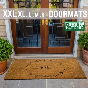 May include: A brown coir doormat with the letter "M" in a wreath design. The doormat is in front of a double wooden door with glass panels. The text "XXL.XL.L.M.s DOORMATS" is at the top of the image. A green sign with the text "NATURAL PLASTIC FREE" is on the right side of the image.