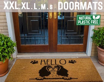 May include: A brown coir doormat with the text "HELLO" and paw prints on either side. A black Cavalier King Charles Spaniel dog is peeking over the top of the mat. The doormat is in front of a wooden door with glass panels. A green leaf with the text "NATURAL PLASTIC FREE" is in the top right corner of the image.