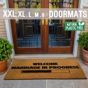 May include: A brown coir doormat with the text "Welcome Marriage in Progress" and a black progress bar underneath. The doormat is placed in front of a wooden door with glass panels. A green leaf sticker with the text "Natural Plastic Free" is on the door.