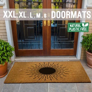 May include: A brown coir doormat with a black sunburst design. The doormat is in front of a wooden door with glass panels. The text "XXL.XL.L.M.s DOORMATS" is above the door. A green sticker with the text "NATURAL PLASTIC FREE" is on the door.