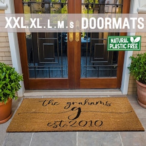 May include: A brown coir doormat with the text "the grahams g est. 2010" in black script font. The doormat is placed in front of a wooden double door with a black metal grid design. A green sign with the text "NATURAL PLASTIC FREE" is visible on the right side of the door.
