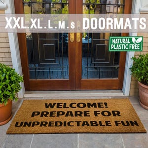 May include: A brown coir doormat with the text "WELCOME! PREPARE FOR UNPREDICTABLE FUN" in black lettering. The doormat is placed in front of a wooden door with glass panels. A green plant is in a terracotta pot to the left of the doormat and another green plant is in a terracotta pot to the right of the doormat. A green sign with the text "NATURAL PLASTIC FREE" is attached to the door.