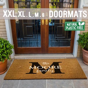May include: A brown coir doormat with the text "the MOORE family" and a large black letter "M" in the centre. The doormat is placed in front of a wooden door with glass panels. A green sign with the text "NATURAL PLASTIC FREE" is visible in the top right corner of the image.