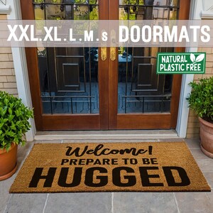 May include: A brown coir doormat with the text "Welcome! Prepare to be Hugged" in black lettering. The doormat is placed in front of a wooden door with glass panels. A green sign above the door reads "Natural Plastic Free".