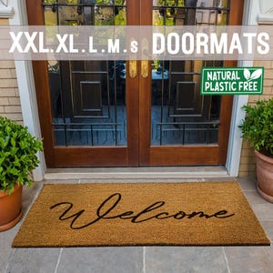 May include: A brown coir doormat with the word "Welcome" written in black cursive script. The doormat is placed in front of a wooden door with glass panels. A green plant is in a terracotta pot to the right of the doormat. The doormat is labelled "Natural Plastic Free".