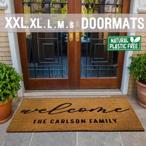 May include: A brown coir doormat with the text "welcome" in a cursive font and "The Carlson Family" in a block font. The doormat is placed in front of a wooden door with glass panels. A green plant is in a terracotta pot to the right of the doormat.
