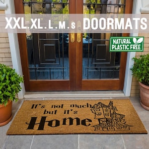 May include: A brown coir doormat with the text "It's not much but it's Home" in black lettering. A black and white illustration of a house with multiple windows is on the right side of the mat. The mat is placed in front of a wooden door with a glass panel.