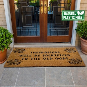 May include: A brown coir doormat with the text "Trespassers will be sacrificed to the old gods" in black lettering. The doormat has a design of stylised birds on each side of the text. The doormat is placed in front of a wooden door with glass panels. A green sign with the text "Natural Plastic Free" is visible in the top right corner of the image.