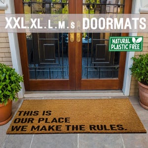 May include: A brown coir doormat with the text "This is our place. We make the rules." in black lettering. The doormat is placed in front of a wooden door with a glass panel. A green leaf sticker with the text "Natural Plastic Free" is on the door.