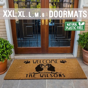 May include: A brown coir doormat with the text "WELCOME" and two paw prints on either side of the text. Below the text is a black silhouette of a Cavalier King Charles Spaniel dog with the text "THE WILSONS" underneath. The doormat is made from natural, plastic-free materials.