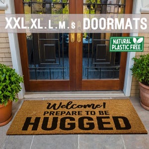 May include: A brown coir doormat with the text "Welcome! Prepare to be Hugged" in black lettering. The doormat is placed in front of a wooden door with glass panels. A green sign with the text "Natural Plastic Free" is visible in the top right corner of the image.