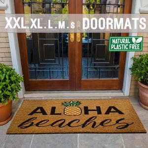 以下が含まれることがあります： 茶色のココナッツ繊維のドアマットに、黒い文字で「ALOHA beaches」と書かれています。黄色のパイナップルに緑の葉が、単語の間に中央に配置されています。画像の右上隅には、「NATURAL PLASTIC FREE」という文字の入った緑色のステッカーがあります。