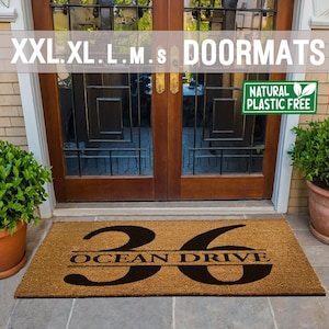 May include: A brown coir doormat with the number 36 and the text "Ocean Drive" in black lettering. The doormat is placed in front of a wooden door with glass panels. A green sign with the text "Natural Plastic Free" is visible in the top right corner of the image.