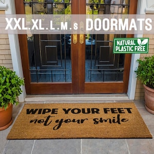 Könnte beinhalten: Eine braune Kokosmatte mit dem Text "Wipe your feet not your smile" in schwarzen Buchstaben. Die Fußmatte befindet sich vor einer Holztür mit Glasscheiben. Ein grünes Schild über der Fußmatte lautet "Natural Plastic Free".