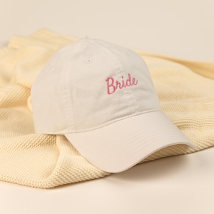 Puede incluir: Gorra de béisbol blanca con visera curva. La palabra "Bride" está bordada en rosa en la parte delantera. La gorra descansa sobre una manta de punto color crema.