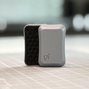 Peut inclure: Un objet rectangulaire en plastique gris aux coins arrondis et à la surface texturée. L'avant présente un petit logo en relief avec les lettres DF. Un composant rectangulaire noir avec une grille d'indentations circulaires est fixé sur le côté.