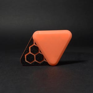 May include: An orange, triangular object rests on a black, triangular base with a honeycomb pattern. The base is textured, and the orange triangle has rounded edges. The background is a solid black.