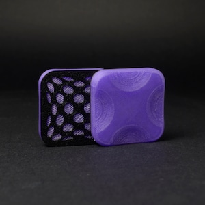 Puede incluir: Dos objetos cuadrados impresos en 3D. Uno es morado liso con patrones curvos, el otro es negro con un patrón de puntos. Ambos tienen esquinas redondeadas y están sobre una superficie negra.