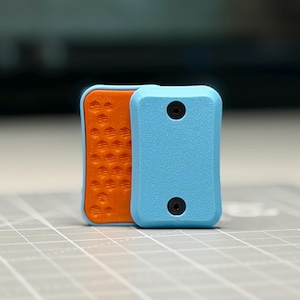 May include: A rectangular fidget toy featuring a light blue textured outer shell with two black circular screws and an orange inner plate with a honeycomb pattern. The object is composed of two distinct parts layered together in a vertical orientation.