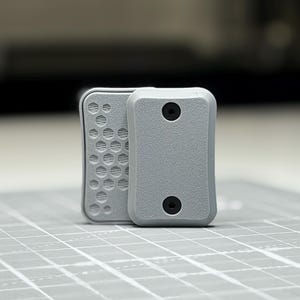 Peut inclure: Deux composants rectangulaires gris clair aux coins arrondis. Une pièce présente un motif en nid d'abeille texturé sur sa surface, tandis que l'autre a une finition granuleuse lisse avec deux vis circulaires noires placées verticalement.