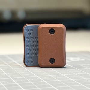 May include: A rectangular object with a textured brown front plate featuring two black hex screws. Behind it, a grey plate is visible, displaying a repeating hexagonal pattern across its surface. The object has rounded corners and a solid, matte finish.