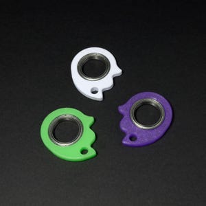 May include: Three colorful fidget spinners in white, green, and purple. Each spinner has a circular bearing in the center and a unique, curved shape with a small hole. The background is black.