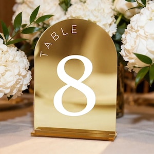 以下が含まれることがあります： 上部に「TABLE」の文字が入った、数字の8のゴールドのテーブルナンバー。テーブルナンバーは、ガラスの花瓶に入った白い花の前にあります。テーブルナンバーは反射素材で作られています。