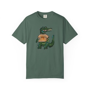 May include: A dark sage green short-sleeve t-shirt featuring a central graphic of a green alligator wearing an orange shirt with the word GATORS printed in black. The illustration has a hand-drawn, textured appearance with visible brush strokes.