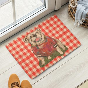 Könnte beinhalten: Eine rechteckige Fußmatte mit einem rot-weiß karierten Muster. Die Matte zeigt eine lächelnde Bulldogge, die einen roten Pullover mit weißen Streifen trägt. Der Hund ist auf einem Stück grünen Grases dargestellt.