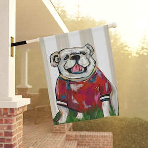 May include: A decorative outdoor flag featuring a smiling bulldog wearing a red and blue shirt. The dog is painted with colorful brushstrokes and stands on green grass against a striped background.