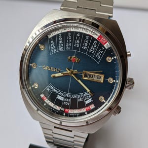 Puede incluir: Un reloj Orient plateado con esfera azul oscuro. El reloj muestra el día, la fecha y el mes. Tiene una correa plateada y una esfera detallada con detalles dorados y marcadores con forma de diamante.