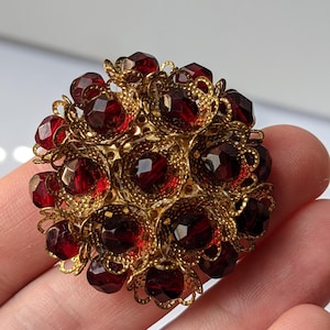 May include: A round brooch featuring a cluster of deep red, faceted, circular glass stones set within an intricate, gold-toned filigree metal frame. The design consists of multiple layers of ornate, lace-like metalwork surrounding each individual stone.
