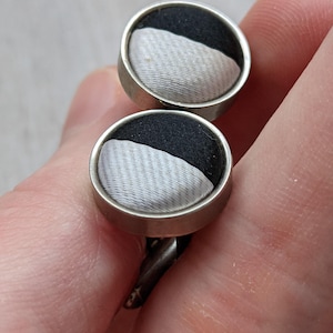 May include: A pair of cufflinks with a silver-coloured metal frame. Each cufflink features a black and white fabric design. The fabric is divided diagonally, with a textured white section and a black section. The cufflinks are held in a hand.