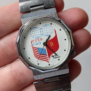 Op de afbeelding: Een vintage polshorloge met een zilverkleurige metalen band en kast. De witte wijzerplaat toont de vlaggen van de VS en de USSR, samen met de tekst "CCCP" en "CWA". Er zijn ook twee wereldbollen afgebeeld.