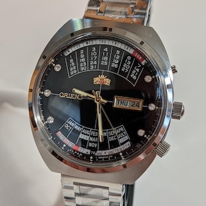 Può includere: Un orologio Orient argentato con quadrante nero, con calendario e visualizzazione del giorno. L'orologio ha un cinturino in acciaio inossidabile e lancette dorate. Il quadrante include indicatori a forma di diamante e la scritta "ORIENT".