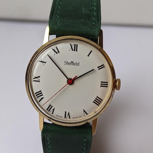 May include: A gold-toned Sheffield wristwatch with a white face, black Roman numerals, and black hands. The watch features a red second hand and a green leather strap. The watch face reads "Sheffield" and "Swiss Made 82".