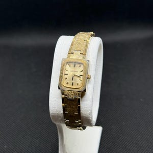 May include: Vintage gold-toned Helbros watch with a square face and matching textured band. The watch is displayed on a white stand against a black background. The watch face has a gold color and the brand name "Helbros" is visible.