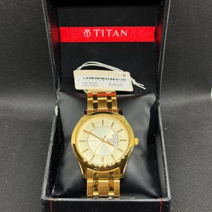 May include: A gold-toned Titan watch with a matching band, displayed in a black box with a red interior. The watch face has a light gold color with gold hands and markers. The box has the Titan logo.