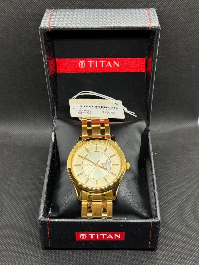 May include: A gold-toned Titan watch with a matching band, displayed in a black box with a red interior. The watch face has a light gold color with gold hands and markers. The box has the Titan logo.