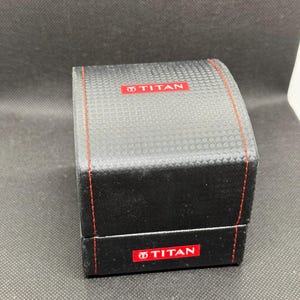 May include: A black watch box with a textured surface and red stitching. The box has the word "TITAN" in red on the top and front. The box is rectangular and has a hinged lid.
