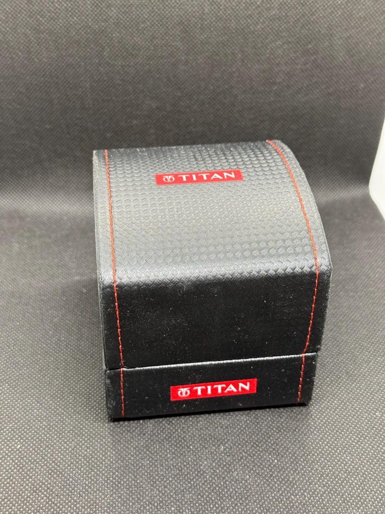 May include: A black watch box with a textured surface and red stitching. The box has the word "TITAN" in red on the top and front. The box is rectangular and has a hinged lid.