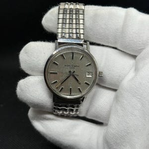 May include: A vintage silver-toned wristwatch with a textured metal band. The watch face is silver with black hour markers and hands. The watch has the text "ETERNA-MATIC 1000" and a date display at the 3 o'clock position. The watch is held by a gloved hand.