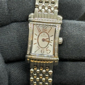 May include: A silver-tone rectangular Bulova wristwatch with a textured center dial, rose gold-tone numerals at 12, 6, and 5, and a matching multi-link metal bracelet. The watch case features small clear stones along the vertical sides.