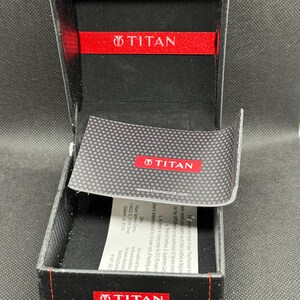 May include: Open black watch box with red accents and the brand name "TITAN" visible on the inside and outside. The box has a carbon fiber pattern on the lid and a small card with text.