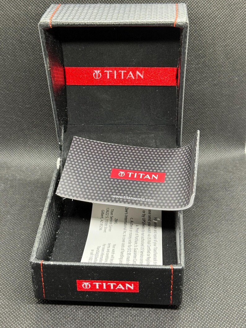 May include: Open black watch box with red accents and the brand name "TITAN" visible on the inside and outside. The box has a carbon fiber pattern on the lid and a small card with text.